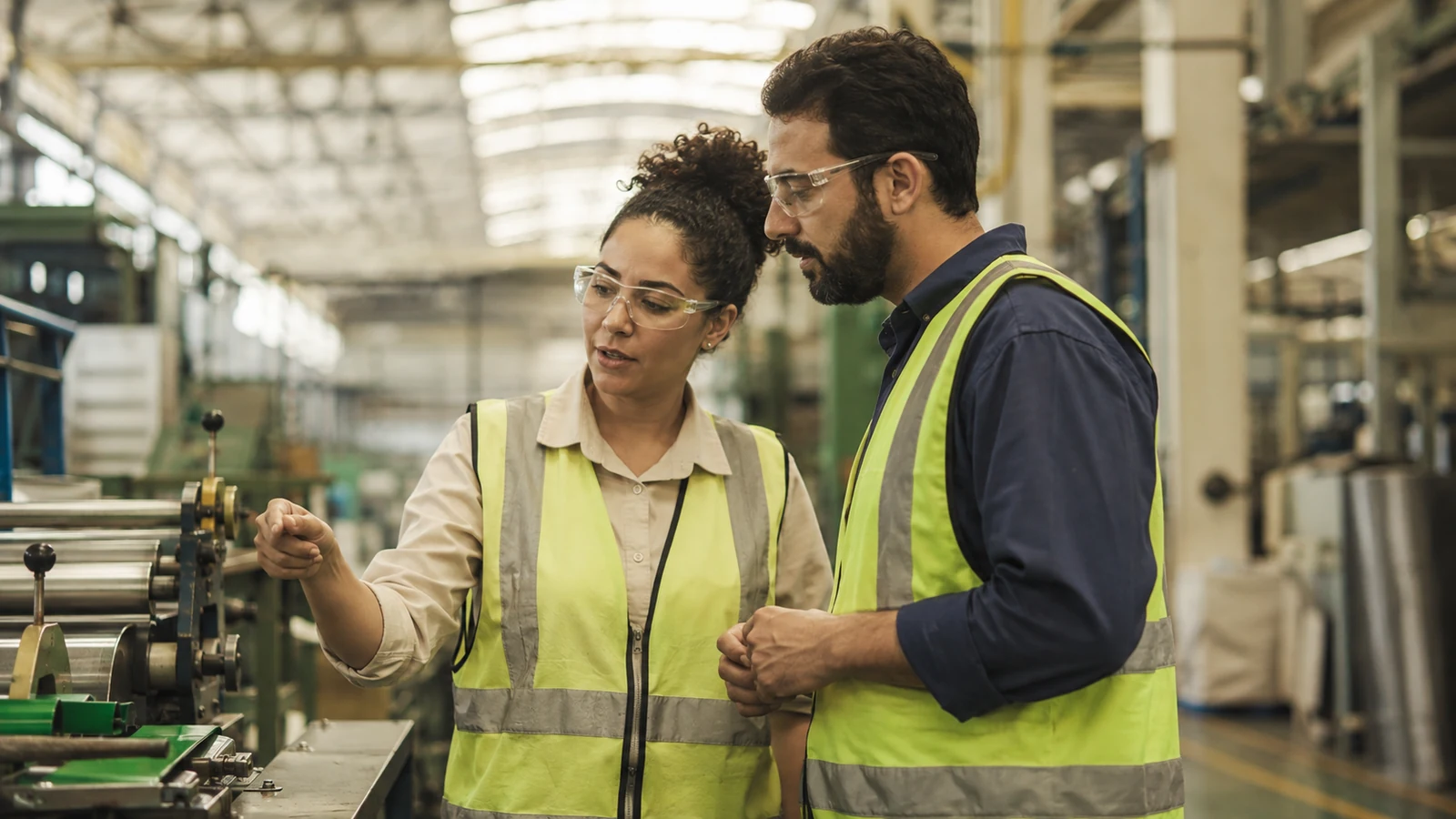 Dois técnicos industriais de óculos de proteção e colete inspecionam uma linha de produção