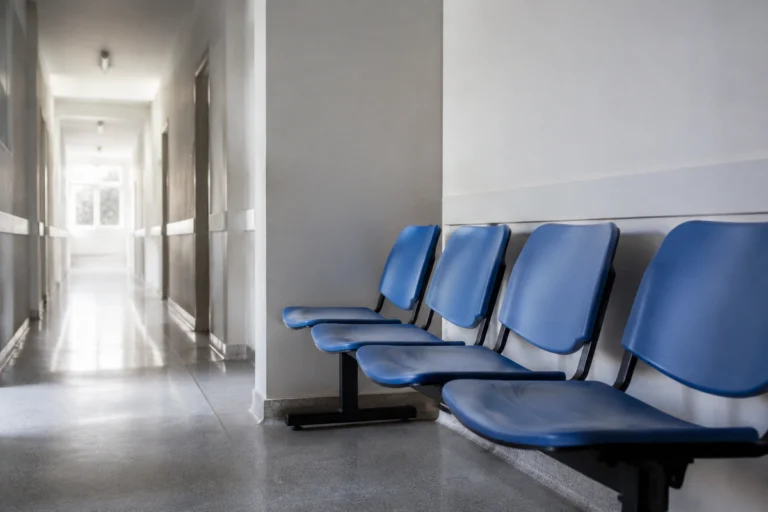 Fileira de cadeiras azuis vazias em sala de espera de unidade de saúde com corredor iluminado