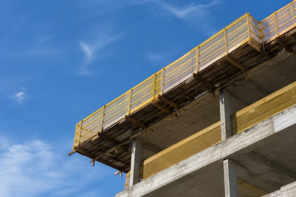 Bandeja de proteção com tela amarela instalada na periferia de edifício em construção, vista de baixo contra o céu