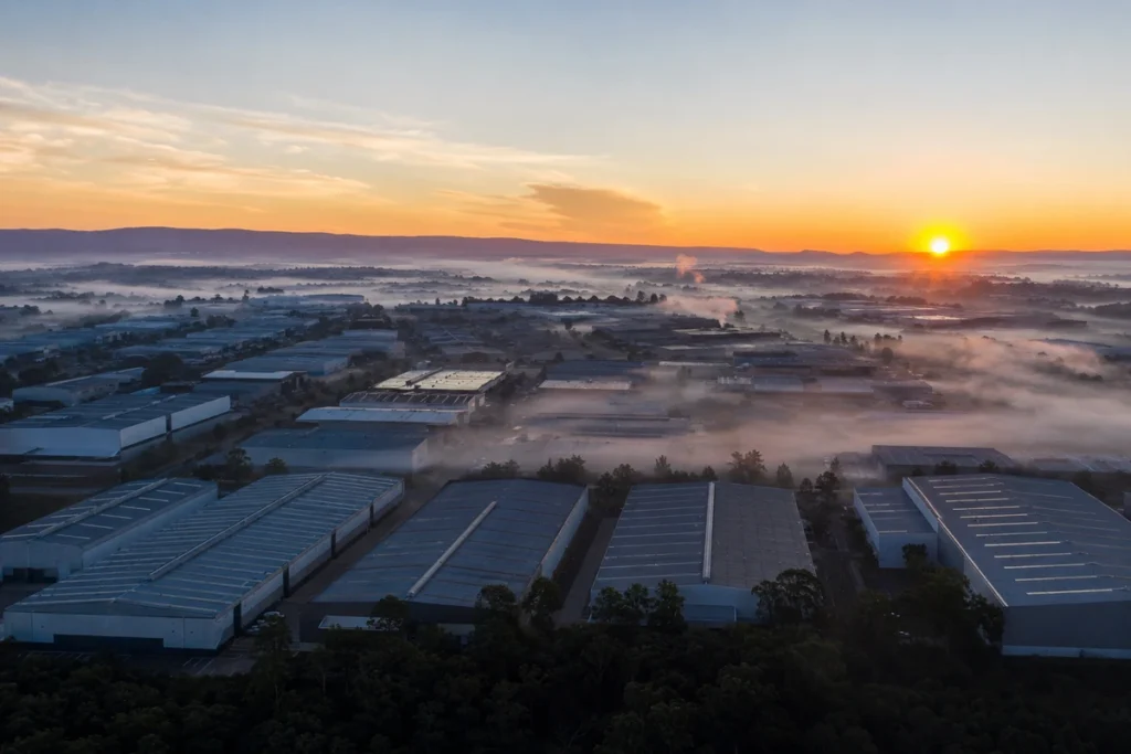 Vista aérea de distrito industrial ao amanhecer com galpões alinhados e neblina baixa entre os telhados